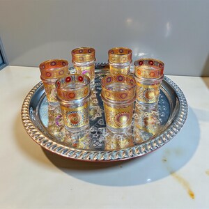 May include: A set of six clear glass tea glasses with gold and colorful decorative patterns. The glasses are arranged on a silver tray with an ornate design. The glasses have a cylindrical shape and are likely used for serving tea or other beverages.