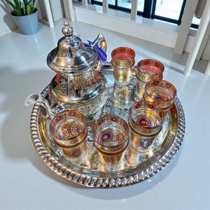 May include: A silver tea set with a teapot, six small glasses, and a serving tray. The glasses and teapot have intricate designs. The glasses are decorated with gold and red patterns. The set is designed for serving tea.