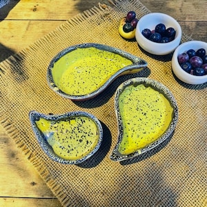 Peut inclure: Trois coupelles en céramique en forme de goutte d'eau avec un intérieur jaune vif parsemé de taches noires et un bord sombre texturé. Deux petits bols blancs remplis de myrtilles fraîches sont placés près des coupelles sur une surface tissée.