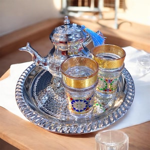 Könnte beinhalten: Ein silbernes Teeservice mit einer Teekanne, zwei Gläsern und einem Tablett. Die Teekanne hat eine blaue Quaste und steht auf einem runden, silbernen Tablett. Die Gläser haben goldene Ränder und bunte Muster. Das Set steht auf einem weißen Tuch.