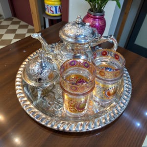 Könnte beinhalten: Ein silbernes Teeservice auf einem dekorativen Tablett. Das Set beinhaltet eine Teekanne, eine Zuckerdose und zwei Gläser. Die Gläser haben bunte Muster. Eine dekorative Vase mit einer Pflanze befindet sich im Hintergrund.