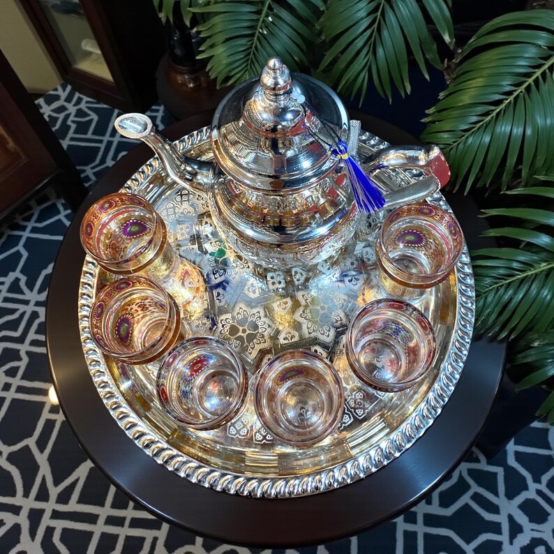 May include: A silver tea set with a teapot, six small glasses, and a serving tray. The teapot has a decorative top and a blue tassel. The glasses and tray have intricate patterns. The set is on a dark wooden table.