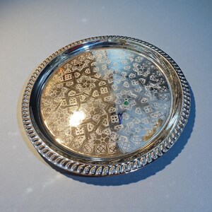 May include: A round, silver-colored tray with an intricate, etched floral pattern on the interior. The tray has a decorative, twisted rope-like edge. A small, green jewel is set within the design.