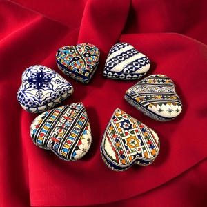 Peut inclure: Six boîtes à bijoux en forme de cœur disposées en cercle. Chaque boîte présente un motif peint à la main unique avec des détails complexes en bleu, jaune, vert, rouge et noir sur fond blanc. Les couvercles arborent des motifs géométriques.