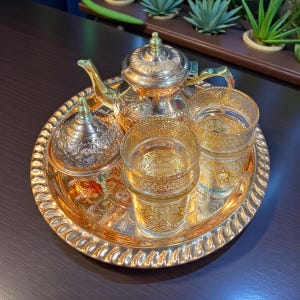 May include: A gold-toned tea set on a matching tray. The set includes a teapot, sugar bowl, and two glasses, all with intricate etched designs. The tray has a decorative, scalloped edge. The set is displayed on a dark surface.