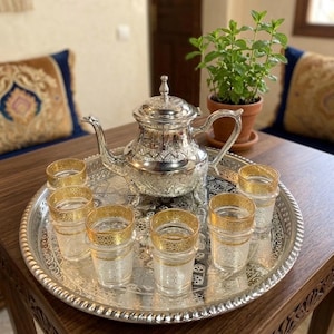 May include: A silver tea set with a decorative teapot and six glasses with gold rims, arranged on a silver tray. A small potted plant sits nearby. The set is on a wooden table, suggesting a tea service.