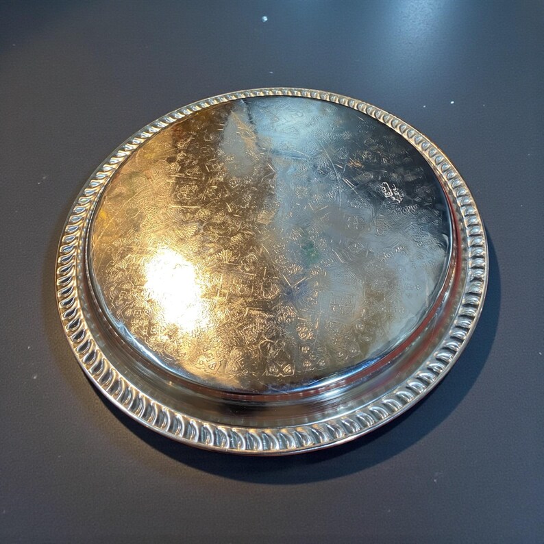 May include: A round, silver-colored tray with a decorative rope-like edge. The tray's surface has a reflective quality and appears to have an embossed floral pattern. The tray is likely made of metal and is suitable for serving or display.