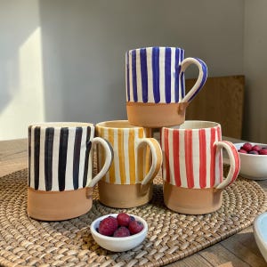 Peut inclure: Quatre mugs en céramique à rayures verticales noires et blanches, jaunes et blanches, rouges et blanches et bleues et blanches. Chaque mug a une base et une anse beiges. Les mugs sont disposés sur un tapis tissé.