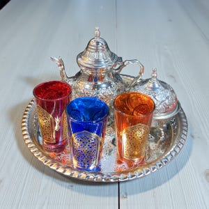 Puede incluir: Un juego de té plateado con tetera, azucarero y bandeja. El juego incluye cuatro vasos coloridos en rojo, azul y naranja, cada uno con diseños dorados. El juego está sobre una superficie de madera clara.