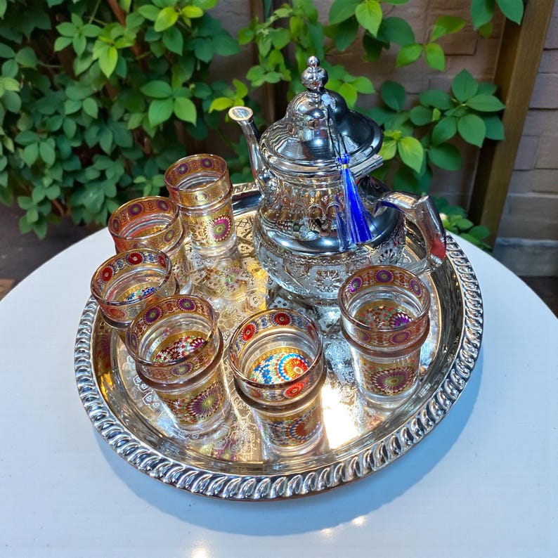 May include: A silver tea set with a teapot, six small glasses, and a serving tray. The glasses and tray have colorful, patterned designs. The teapot has a blue tassel and is surrounded by green foliage.