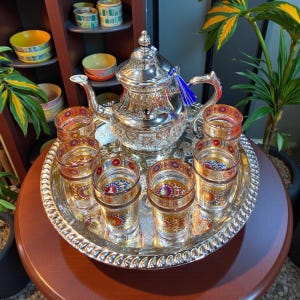 Könnte beinhalten: Ein silbernes Teeservice mit einer Teekanne, einem Tablett und sechs Gläsern. Die Teekanne hat aufwendige Designs und eine blaue Quaste. Die Gläser und das Tablett haben farbenfrohe Muster. Das Set steht auf einem runden Holztisch.