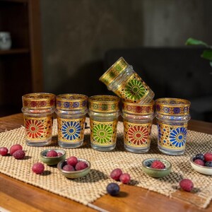 Op de afbeelding: Een verzameling van zes decoratieve glazen theekopjes met gouden randen en kleurrijke geometrische ontwerpen in rood, blauw en groen. De kopjes zijn gerangschikt op een geweven mat met kleine kommen met bessen.