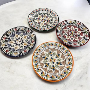 Puede incluir: Cuatro platos decorativos de cerámica con coloridos patrones geométricos. Cada plato presenta un diseño central en forma de estrella en varios colores, incluyendo azul, verde, naranja y rojo, sobre un fondo blanco. Los platos tienen forma redonda y están dispuestos sobre una superficie clara.