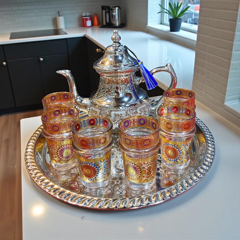 May include: A silver tea set with a teapot and several small glasses on a decorative tray. The glasses and teapot are adorned with colorful patterns. A blue tassel hangs from the teapot's handle.