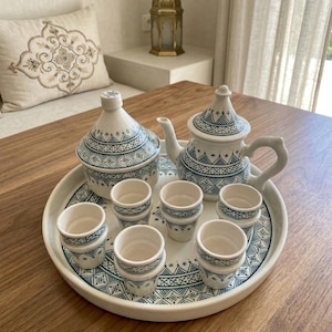 Op de afbeelding: Een witte keramische theeset met blauwe geometrische patronen. De set bevat een theepot, een suikerpot en zes kleine kopjes, allemaal gerangschikt op een bijpassend dienblad. De diameter van het dienblad is ongeveer 30 cm.