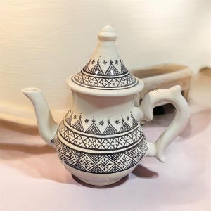 Puede incluir: Tetera de cerámica blanca con patrones geométricos negros. La tetera presenta una tapa cónica, un asa curva y un pico. El diseño incluye triángulos, cruces y otras formas. La tetera se utiliza probablemente para servir té.