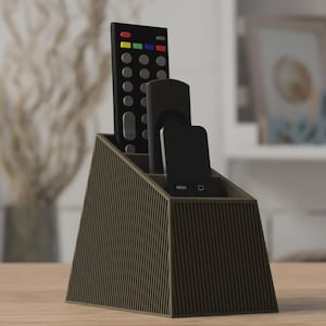 Peut inclure: Un support de télécommande trapézoïdal marron foncé avec une texture nervurée verticale. L'organisateur comporte trois compartiments étagés de hauteurs différentes, contenant actuellement une grande télécommande et deux petites télécommandes.