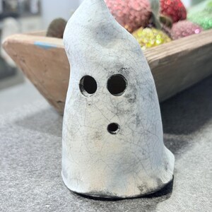 May include: A ceramic ghost figurine with a distressed, crackled finish. The ghost has two large eye holes and a smaller mouth hole. The figurine is light gray with darker gray accents and sits on a gray surface. The ghost is a Halloween or spooky-themed decorative item.