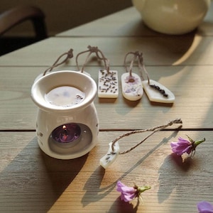 Op de afbeelding: Een witte keramische etherische olieverspreider met een brandende paarse kaars, geurwasmelts en een theepot op een houten oppervlak. De wasmelts zijn rechthoekig en ovaal van vorm, met bloemen en botanische ontwerpen.