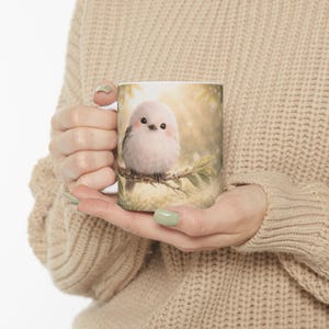 May include: White ceramic mug featuring a charming illustration of a fluffy, light pink bird perched on a branch. The bird has black eyes and a small beak. The background is a soft, blurred image of a forest scene with sunlight.