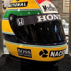 以下が含まれることがあります： 黄色いレーシングヘルメット。緑と青の水平ストライプ、Hondaロゴ入りのダークティンテッドバイザー、Marlboro、National、Bossなどのスポンサーデカールが特徴。台座の金属プレートには「Ayrton Senna 1988 Replica Helmet」と刻印されている。