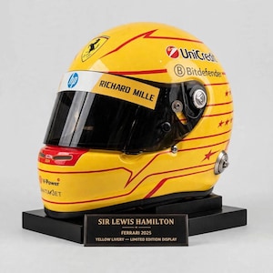 May include: A yellow racing helmet featuring red horizontal stripes and star accents. The visor strip displays the text HP and RICHARD MILLE. Logos for Ferrari, UniCredit, Bitdefender, and V-Power are visible. A plaque reads SIR LEWIS HAMILTON FERRARI 2025.