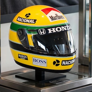 May include: A yellow racing helmet featuring green and black horizontal stripes, a clear visor, and various sponsor logos including Nacional, Marlboro, Honda, Rheos, and Boss. The helmet is displayed on a black stand within a glass enclosure.