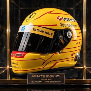 Op de afbeelding: Een gele racehelm met rode horizontale strepen en sterren, voorzien van logo's van Ferrari, HP, Richard Mille, UniCredit, Bitdefender en V-Power. Een zwart displayplaatje vermeldt: SIR LEWIS HAMILTON FERRARI 2025 YELLOW LIVERY.