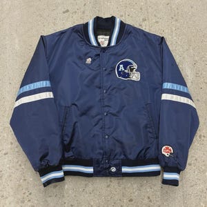May include: Navy blue satin bomber jacket with light blue and white stripes on the sleeves and hem. Features a football helmet logo with the letter "A" and a small emblem on the chest. A vintage sports jacket.
