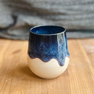Pode incluir: Um copo de cerâmica artesanal com um design de duas cores. A metade superior é azul escuro com um esmalte texturizado, passando para uma base branca cremosa. A borda tem um detalhe marrom escuro. O copo está sobre uma superfície de madeira.