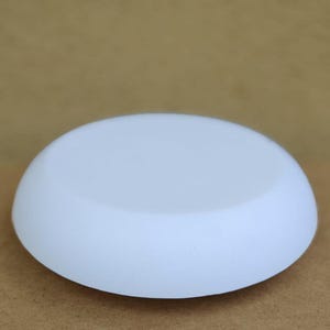 Op de afbeelding: Een wit, afgerond, schijfvormig object met een glad oppervlak. Het object heeft een effen, ondoorzichtige witte kleur en lijkt te zijn gemaakt van een niet-reflecterend materiaal. Het object ligt op een bruin oppervlak.