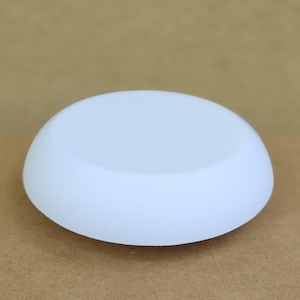 May include: A white, smooth, rounded object with a shallow, concave top. The object is a solid, opaque white and sits on a light brown surface. The shape is reminiscent of a small, flattened bowl or a smooth stone.