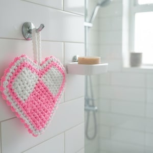 Può includere: Una spugna da bagno a forma di cuore, rosa e bianca, appesa a un gancio d'argento. La spugna ha un motivo a scacchiera e un anello bianco per appenderla. Lo sfondo è un bagno piastrellato di bianco.