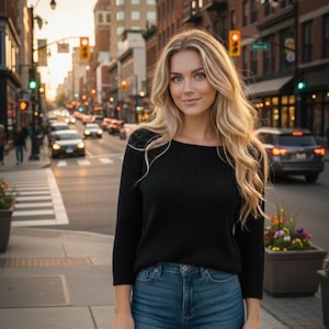 Puede incluir: Una mujer con cabello rubio largo está de pie en una calle de la ciudad. Lleva una camisa negra de manga larga y vaqueros azules. El fondo muestra edificios, tráfico y un paso de peatones.