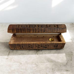 Può includere: Scatola per incenso in legno aperta, di colore marrone caldo. Il coperchio e i lati sono decorati con motivi floreali scolpiti. All'interno è visibile un piccolo supporto rotondo color ottone.