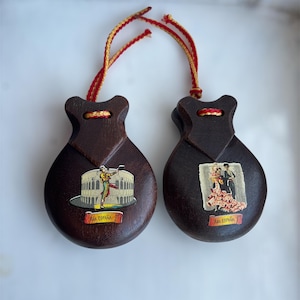 May include: A pair of dark brown wooden castanets connected by red and yellow braided cords. Each castanet features a decorative decal with the text "Recuerdo ESPAÑA". One shows a bullfighter in an arena, and the other shows a flamenco dancer.