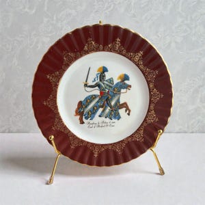 Op de afbeelding: Een decoratieve vintage bord met een bordeauxrode rand en gouden details. Het midden toont een ridder te paard in blauw, geel en wit. Het bord wordt gepresenteerd op een gouden standaard, met een historisch ontwerp.