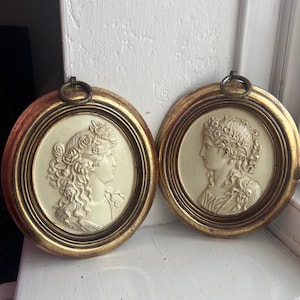 Puede incluir: Dos placas de pared ovaladas antiguas. Cada placa presenta un perfil en bajorrelieve crema de una mujer con peinados ornamentados, enmarcado en un marco dorado. Los marcos tienen un anillo decorativo en la parte superior para colgar.