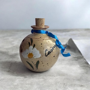 May include: A round, speckled beige ceramic bottle with a cork stopper. The bottle features a hand-painted white daisy with a yellow centre and green leaves. The word "Cassis" is written in black script. A bright blue ribbon is tied around the neck.