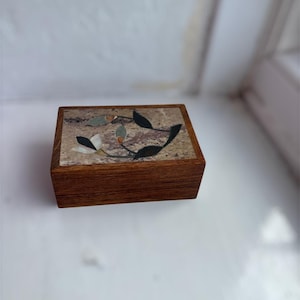 May include: A rectangular wooden box with a hinged lid featuring an inlaid stone panel. The inlay displays a floral motif with white, black, and grey stone petals and leaves set against a mottled beige and brown stone background.