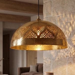 Pode incluir: Uma luminária pendente em forma de cúpula feita de latão polido, com padrões intrincados cortados a laser que projetam sombras detalhadas na parede de pedra adjacente. A lâmpada pende de um fio preto fino, emitindo um brilho dourado quente.