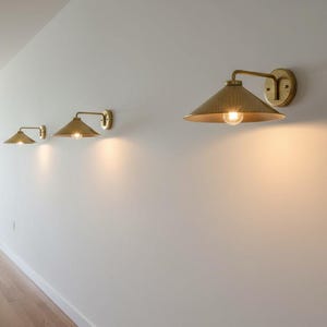 May include: Three brass wall sconces with conical shades are mounted on a white wall. The sconces are arranged in a row, each with a warm light emanating from the bulb. The hallway has a wooden floor and a vase with greenery.