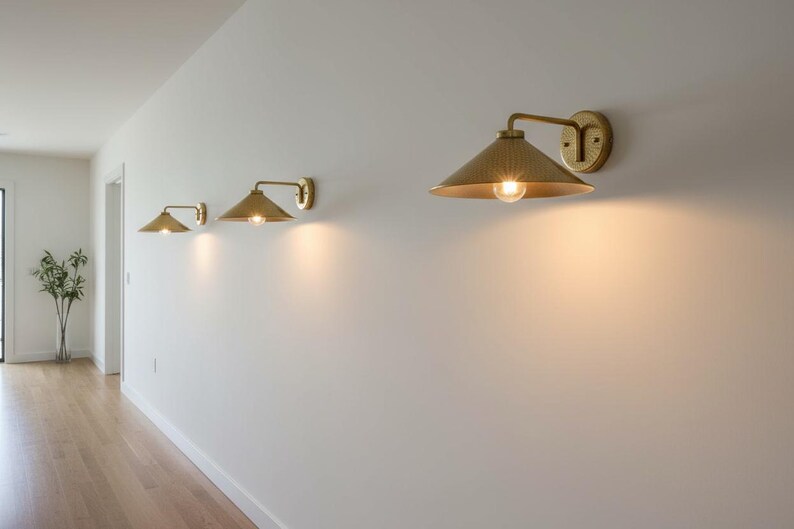 May include: Three brass wall sconces with conical shades are mounted on a white wall. The sconces are arranged in a row, each with a warm light emanating from the bulb. The hallway has a wooden floor and a vase with greenery.