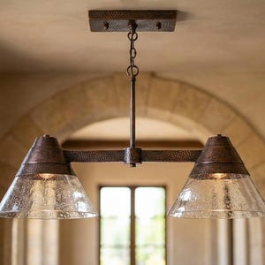 May include: A dual-light pendant fixture featuring a dark, hammered copper finish on the metal frame and canopy. Two conical, seeded glass shades hang from a horizontal bar, suspended by a central rod and a short chain from the ceiling mount.