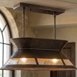 May include: A rectangular pendant light fixture with a dark bronze, hammered metal finish. The fixture features two tiers of flared metal shades and a central band of textured, seeded glass panels that allow light to emit from two internal bulbs.