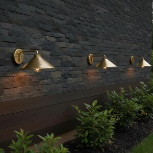 May include: Outdoor wall-mounted lights with a gold finish. The lights are mounted on a dark gray stone wall. The lights have a conical shade and are positioned above a dark wood panel. Green bushes are planted below the lights.