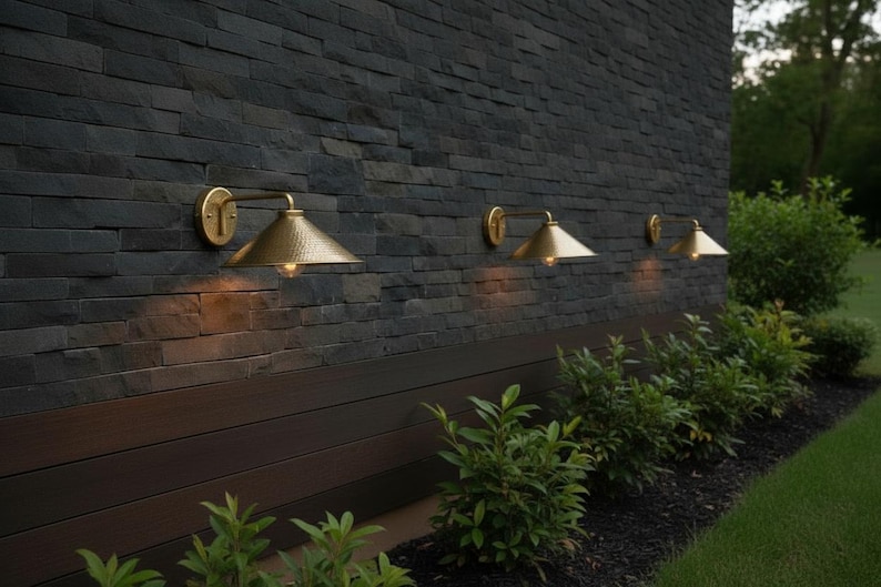 May include: Outdoor wall-mounted lights with a gold finish. The lights are mounted on a dark gray stone wall. The lights have a conical shade and are positioned above a dark wood panel. Green bushes are planted below the lights.