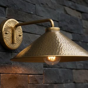 May include: A brass wall sconce with a textured, cone-shaped shade and a round backplate. The sconce is mounted on a dark gray stone wall. The light bulb is visible inside the shade.