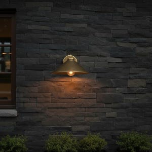 May include: A brass outdoor wall-mounted light fixture with a conical shade and a single exposed bulb. The light is mounted on a dark gray stone wall. The warm light contrasts with the cool tones of the wall.