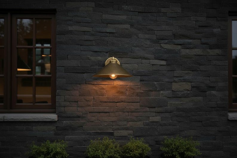 May include: A brass outdoor wall-mounted light fixture with a conical shade and a single exposed bulb. The light is mounted on a dark gray stone wall. The warm light contrasts with the cool tones of the wall.
